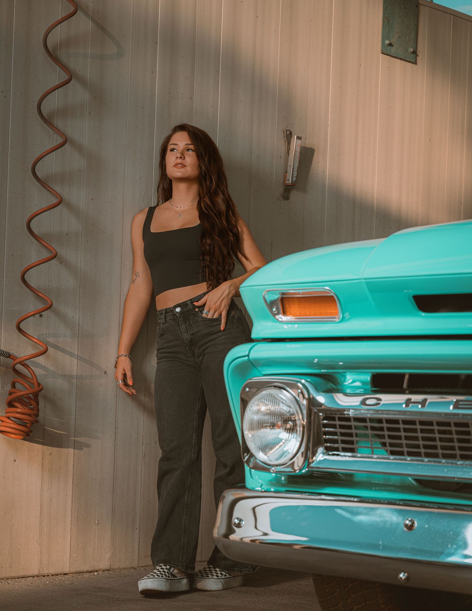 Young woman in black crop top and jeans posing beside a classic turquoise Chevrolet truck in a garage.