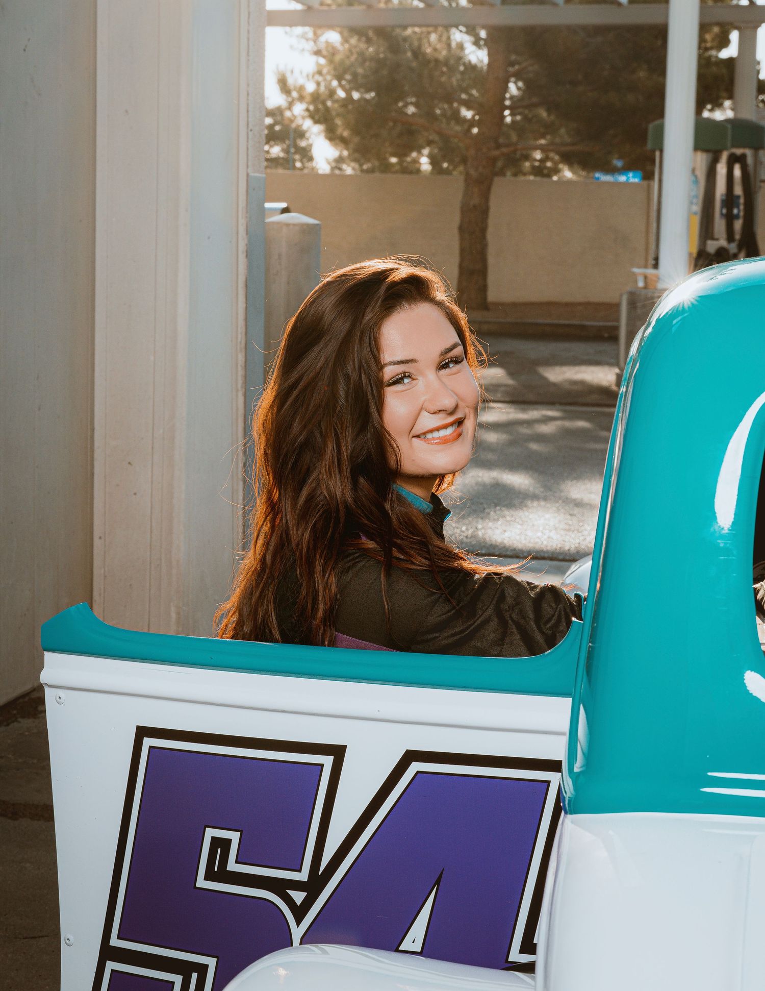 Young woman with long brown hair smiling while sitting in a teal and white vintage race car numbered 54.