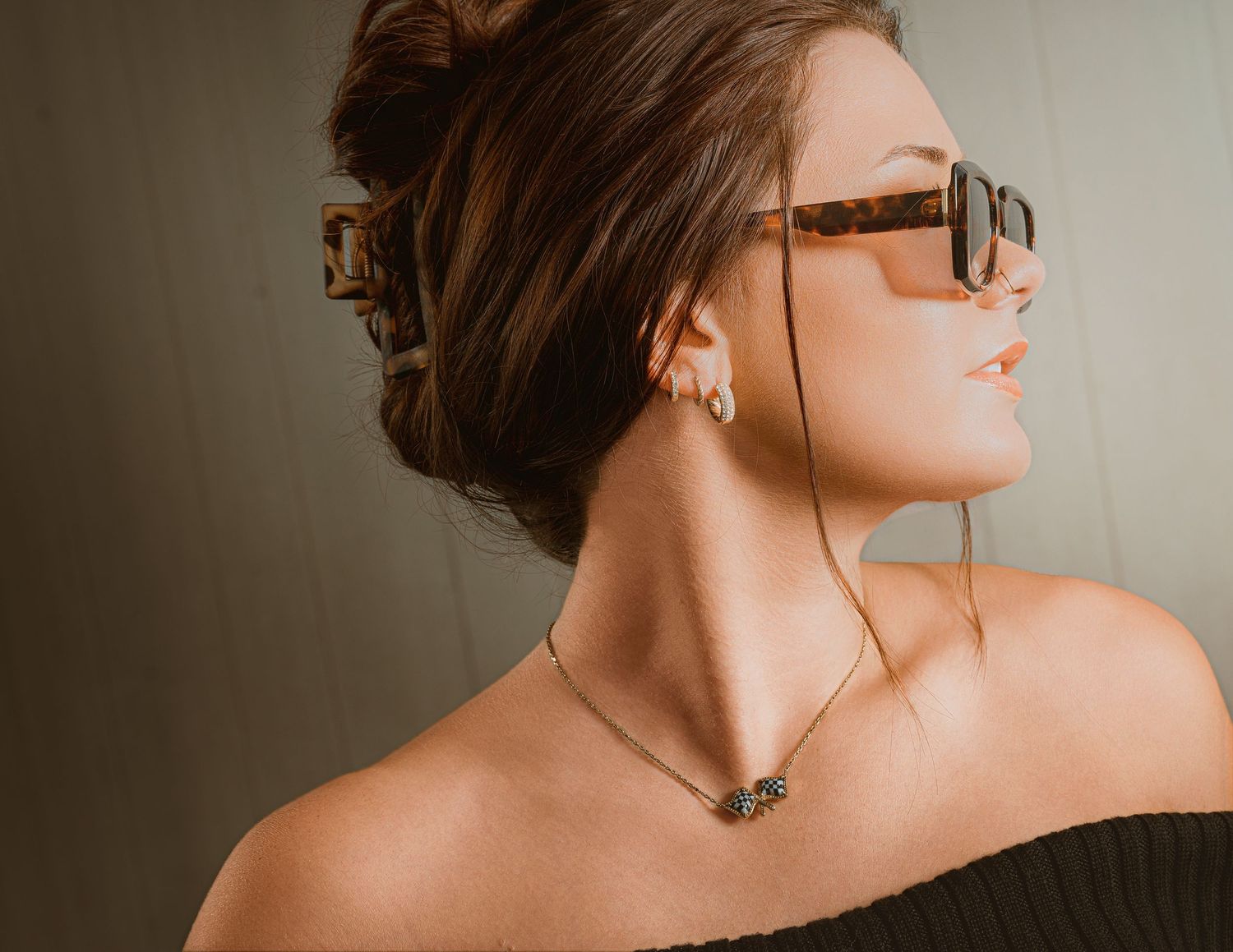 Woman in tortoiseshell sunglasses and gold necklace with updo hairstyle wearing off-shoulder top.