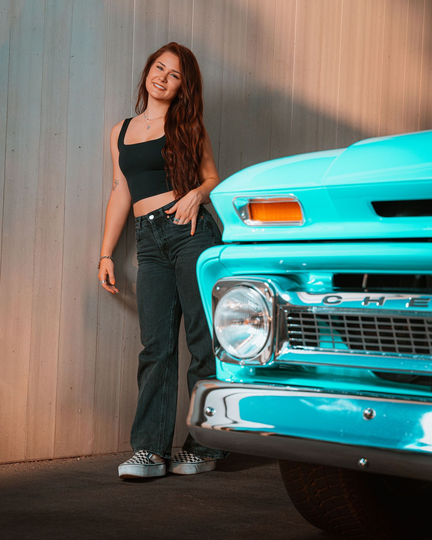 Young woman in black crop top and jeans posing beside a classic turquoise Chevrolet truck against a metal wall.