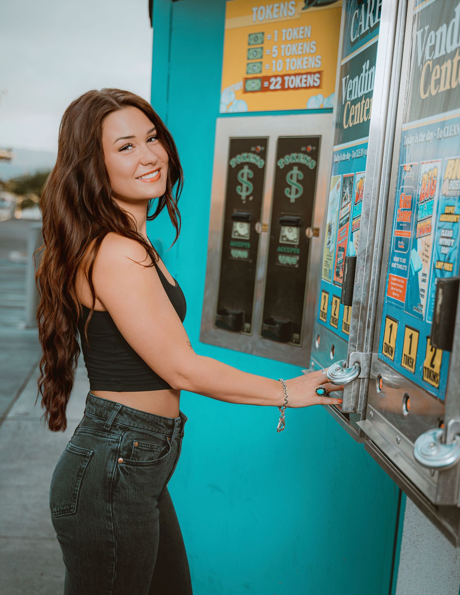 A smiling brunette woman in a black crop top and jeans poses at a teal token vending machine outdoors.