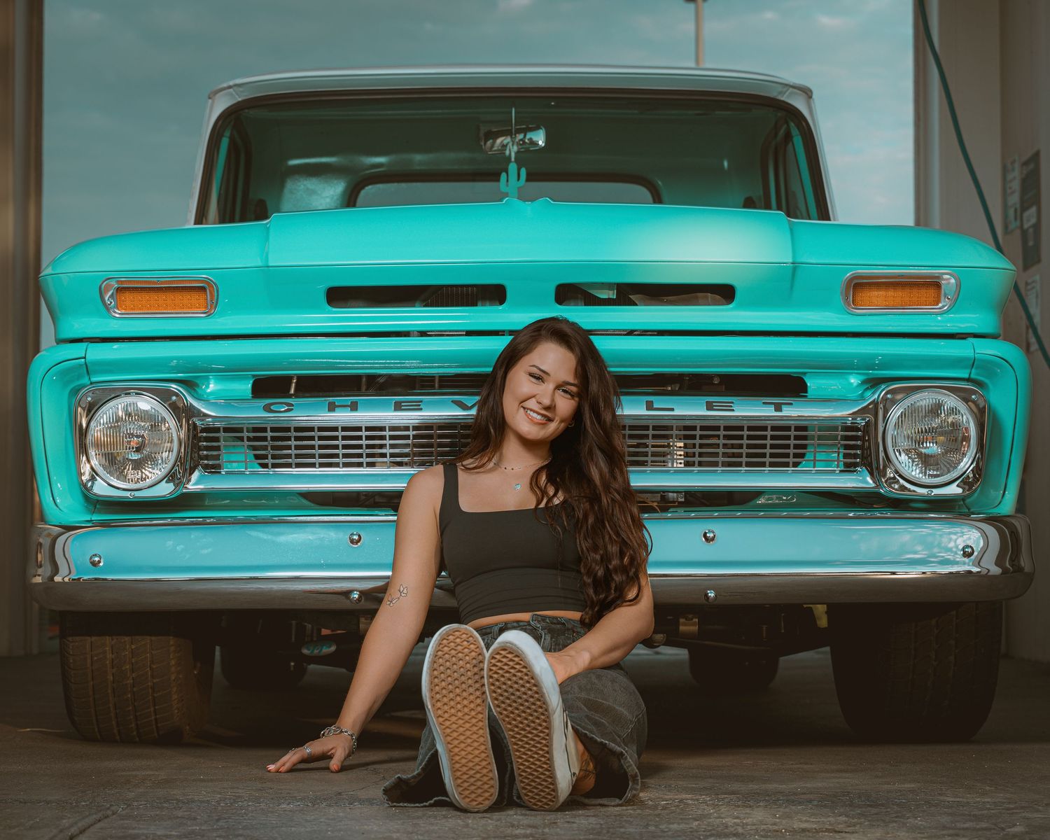 Young woman sitting in front of a classic turquoise Chevrolet pickup truck in a garage setting.