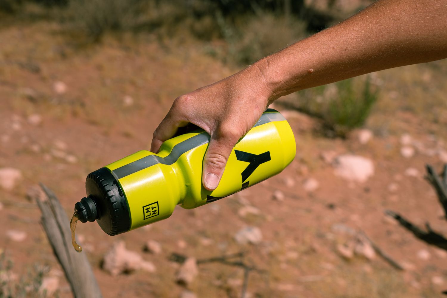Hand holding yellow LMNT branded sports water bottle outdoors in desert trail environment.