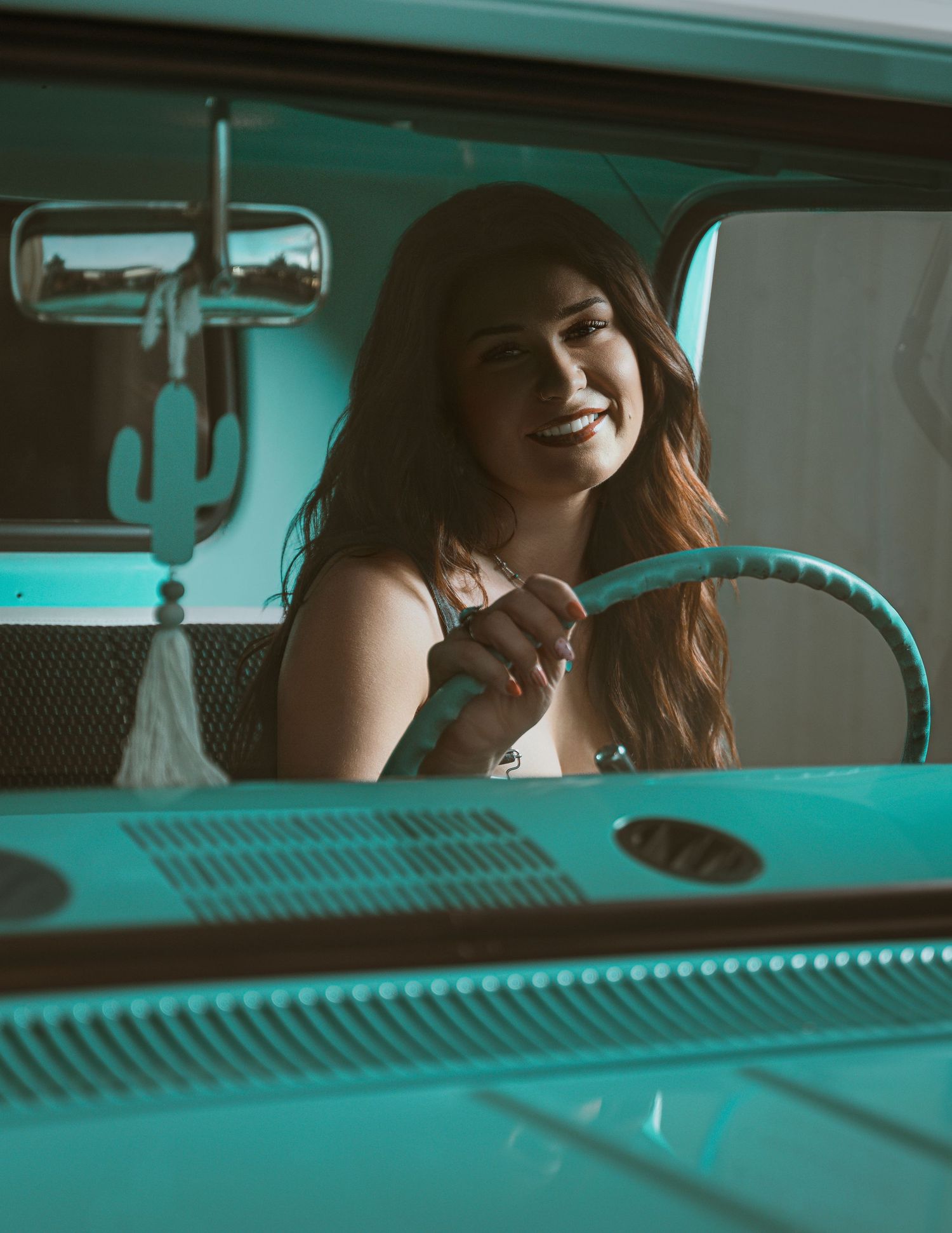 Smiling woman with long brown hair sitting in a vintage turquoise VW bus holding the steering wheel.