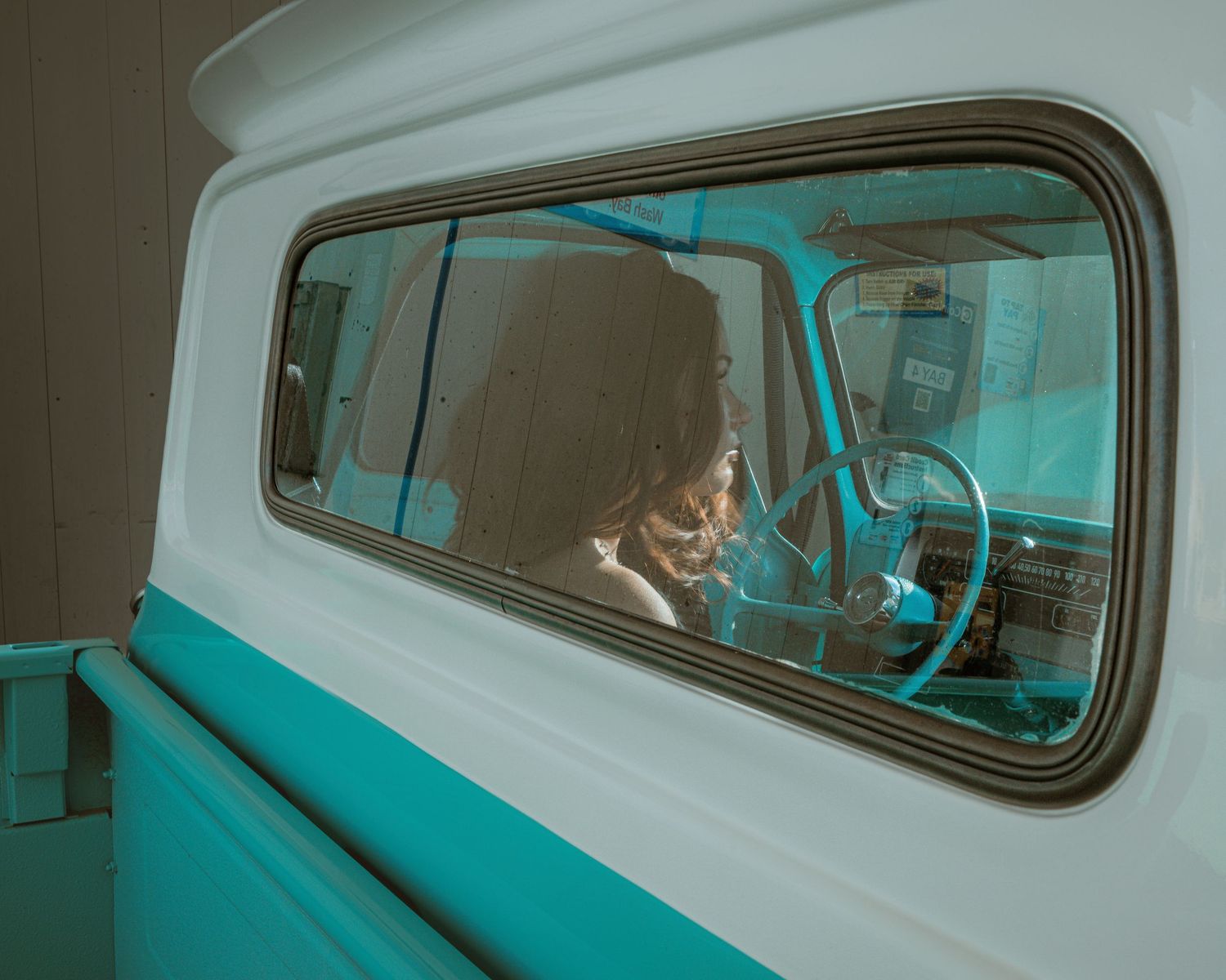 A person sits inside a vintage teal and white truck, visible through the rear window in soft natural light.