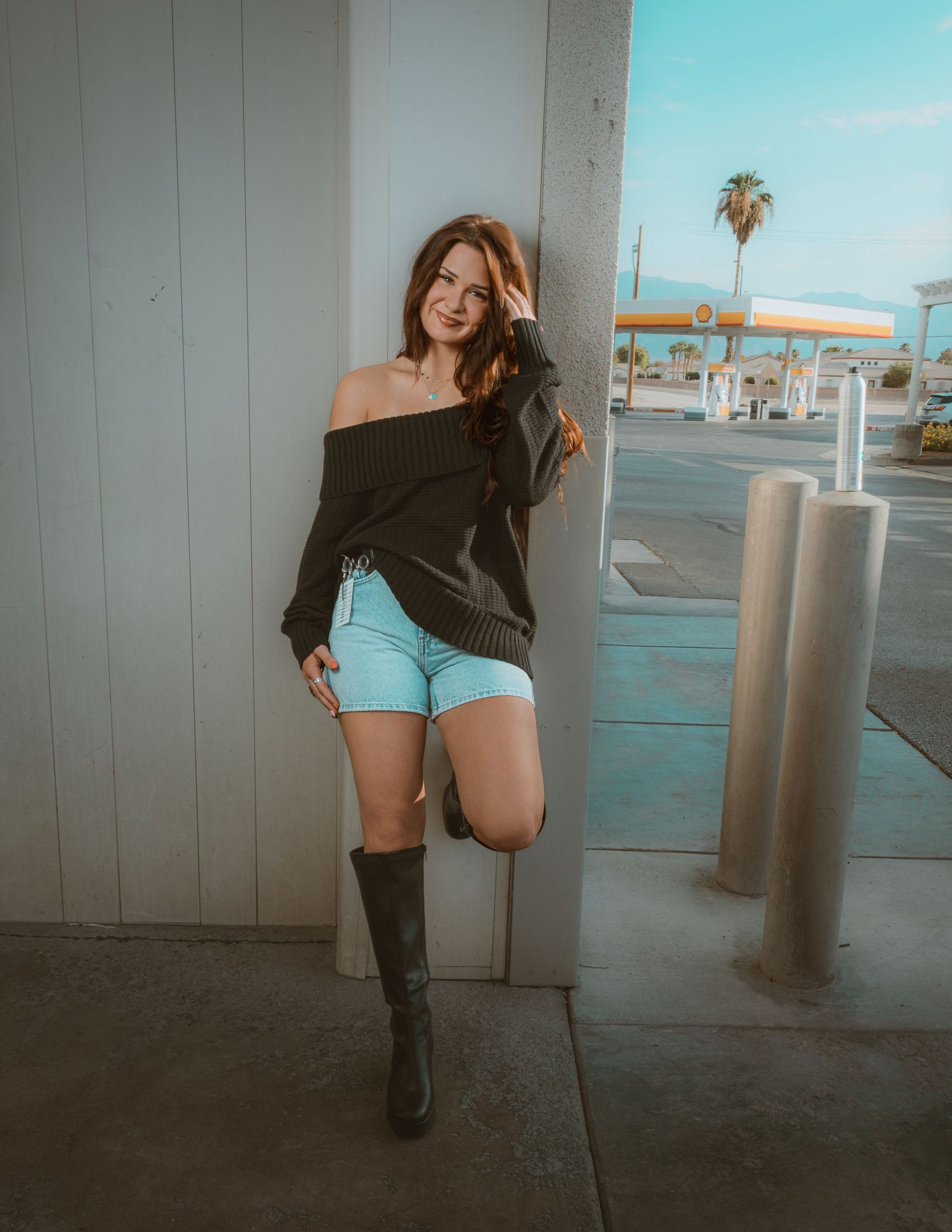 Young woman in black off-shoulder sweater, denim shorts, and knee-high boots posing by a wall.