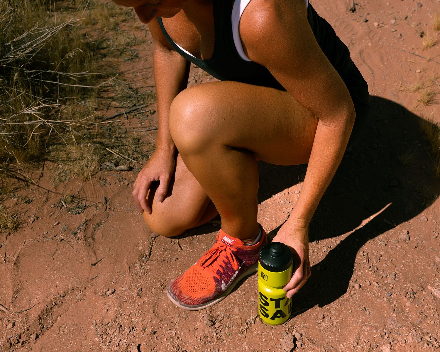 Female trail runner crouching on red dirt trail holding a yellow LMNT water bottle wearing orange sneakers.