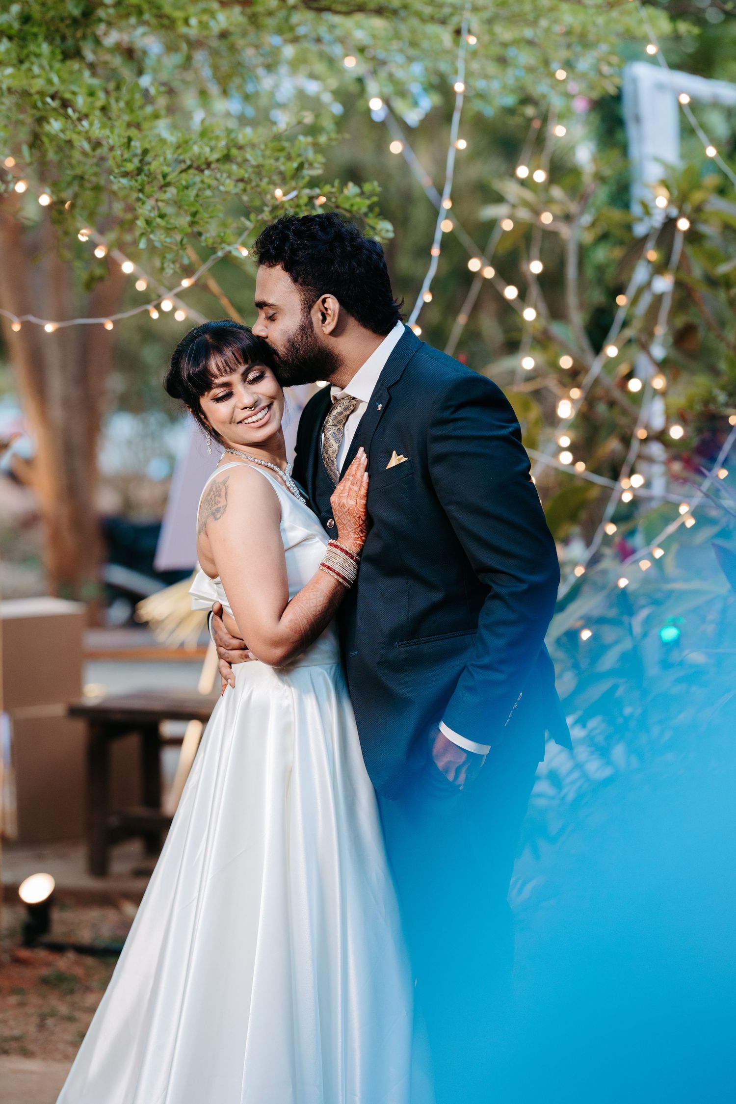 Bride in white gown and groom in dark suit share a romantic moment under string lights at an outdoor wedding.