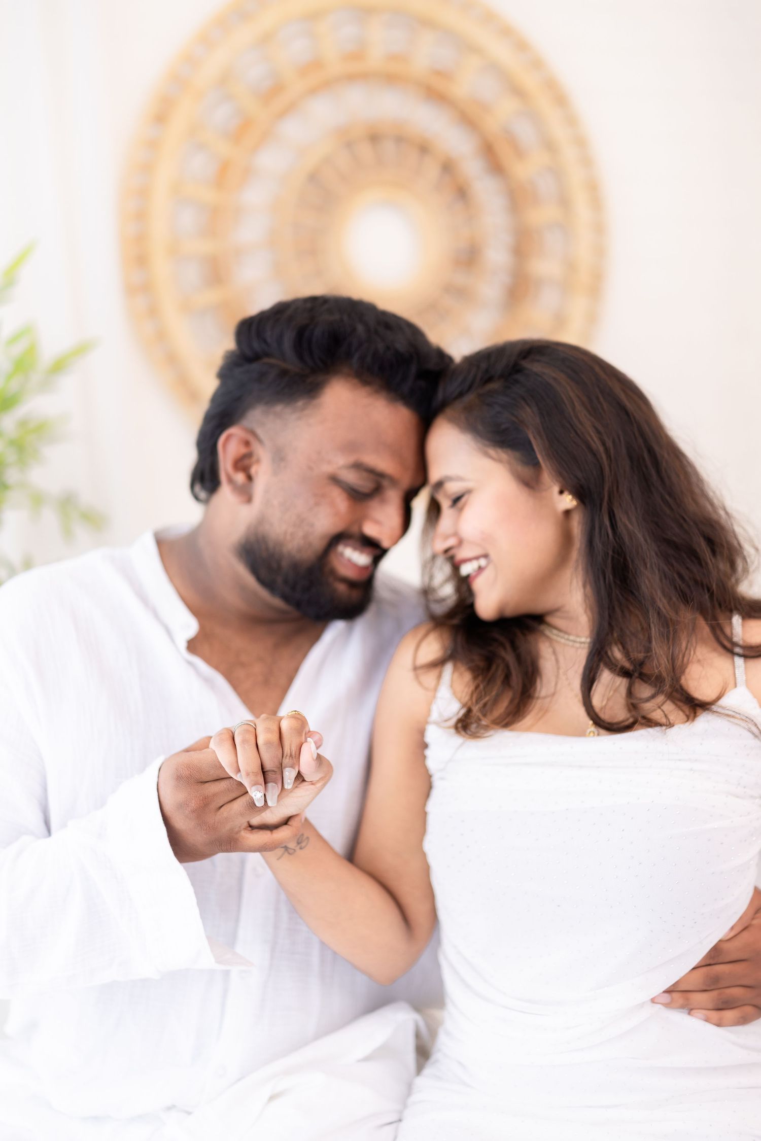 Happy Indian couple in white outfits sharing a tender moment, smiling and holding hands in a bright, elegant room.