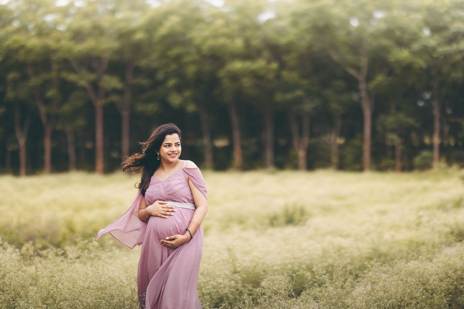Pregnant woman in flowing lavender gown standing in a meadow, smiling warmly with trees in background.
