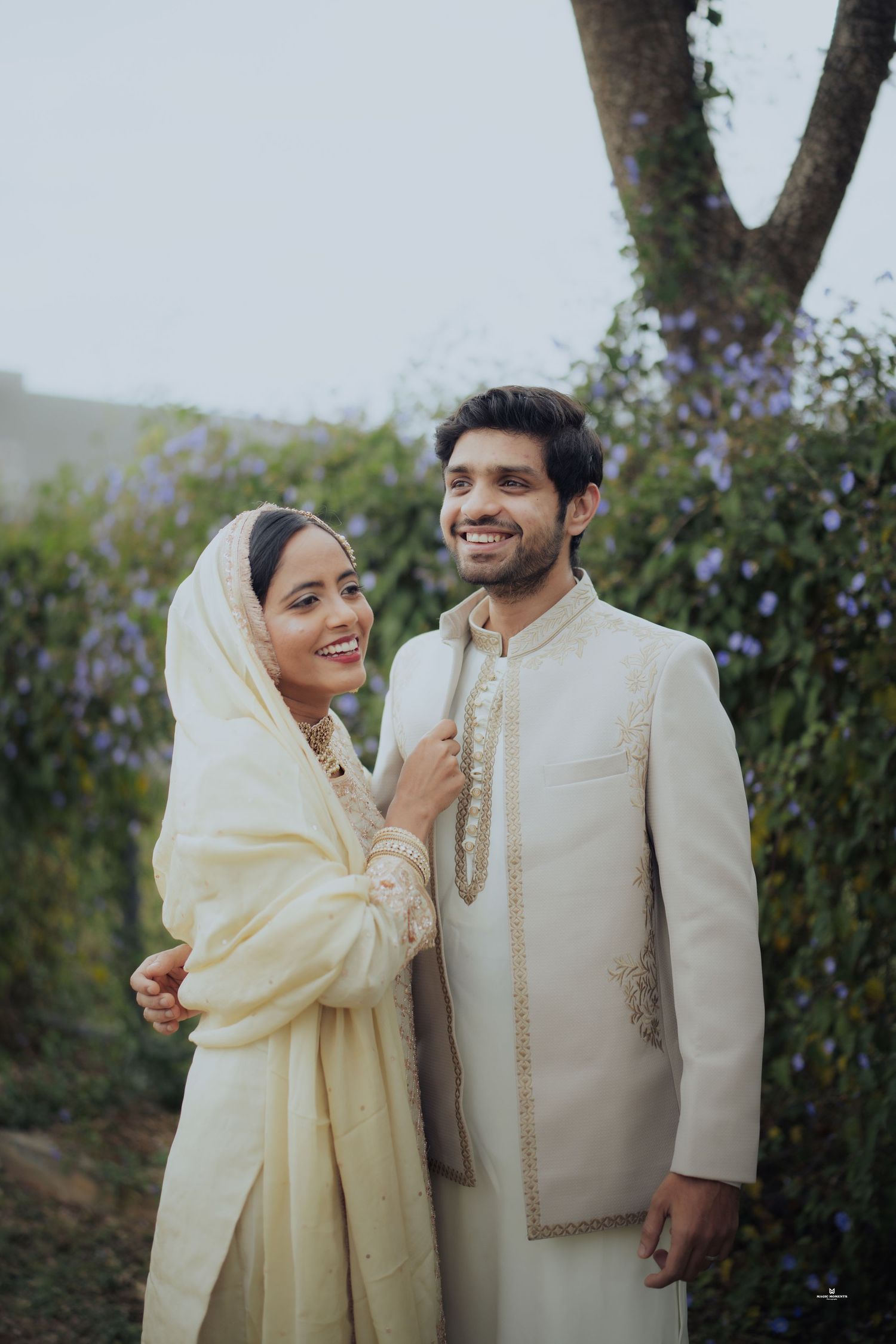 Couple in matching cream traditional Indian wedding attire posing outdoors with lush greenery backdrop.