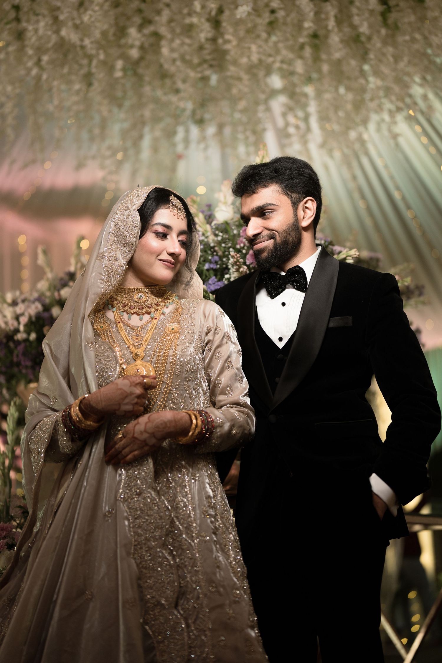 A bride in gold bridal attire and groom in black tuxedo pose elegantly at a floral wedding reception.
