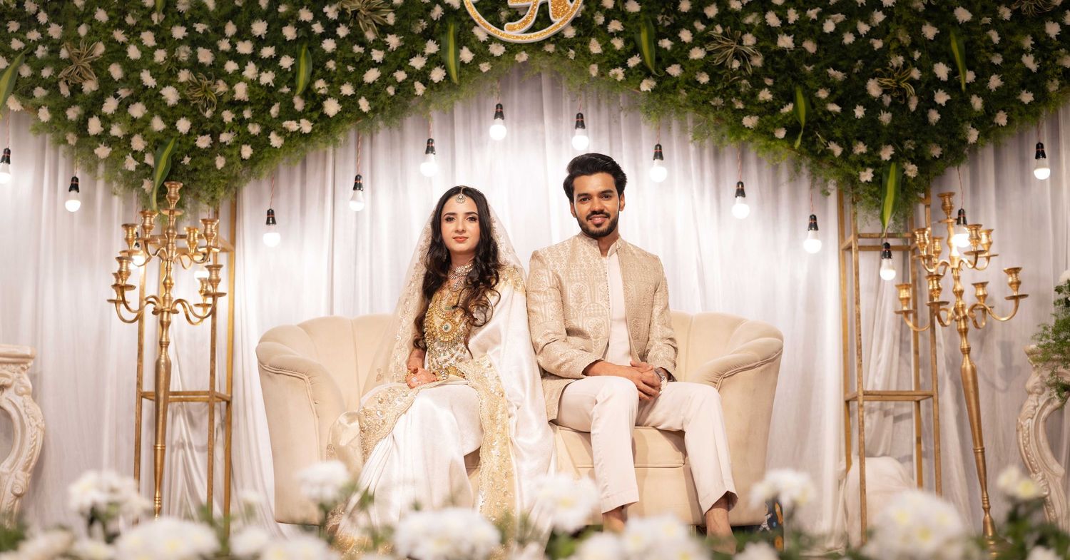 Elegant South Asian couple in traditional wedding attire seated on white sofa under floral arch with white blooms and greenery.