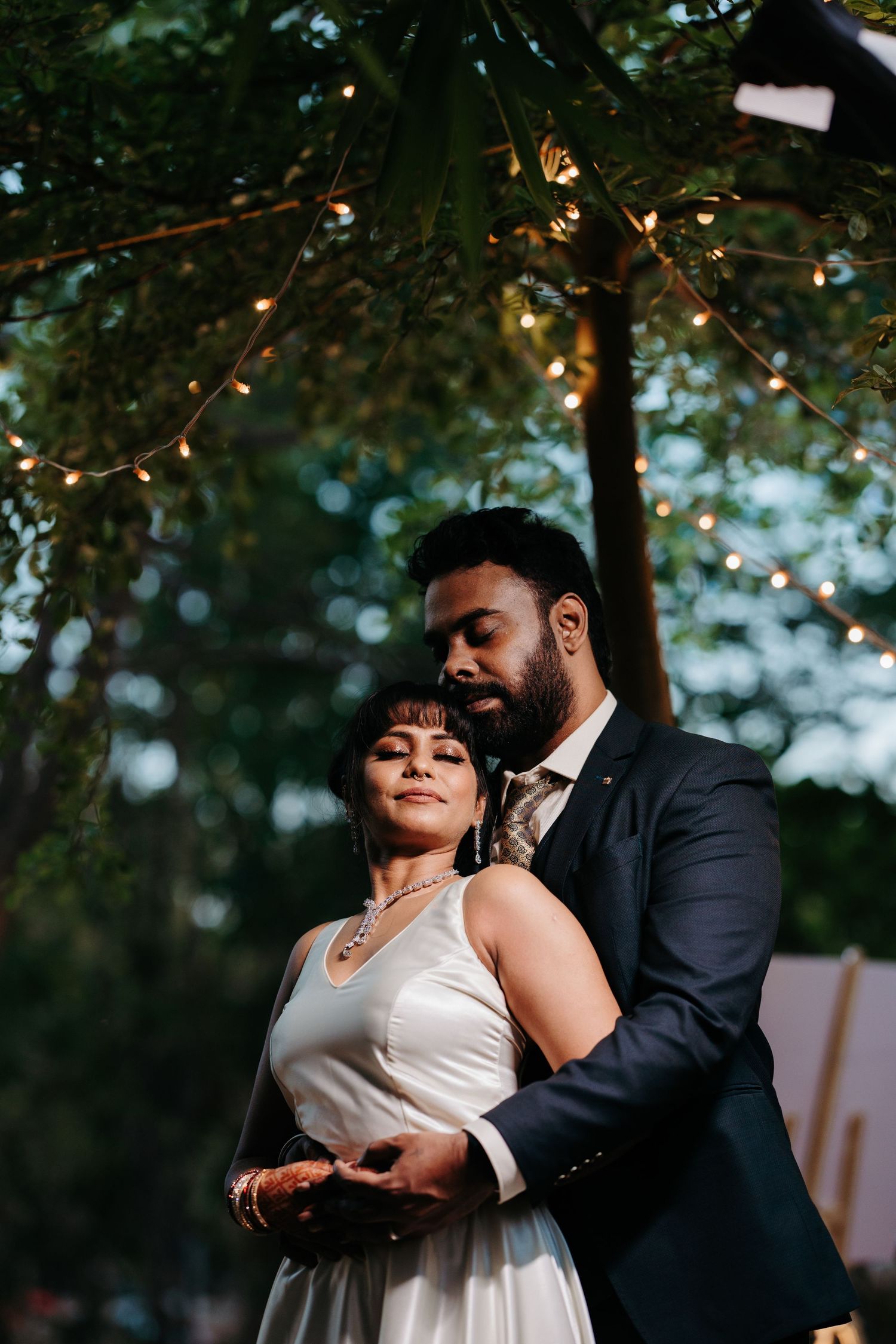 Couple sharing a romantic embrace outdoors at dusk, bride in white dress, groom in dark suit, string lights above.