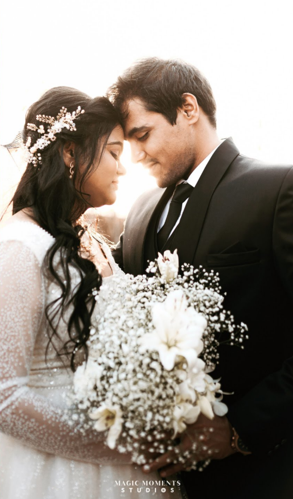 A bride and groom share an intimate moment, she holds a white floral bouquet, both dressed in wedding attire with soft backlight.
