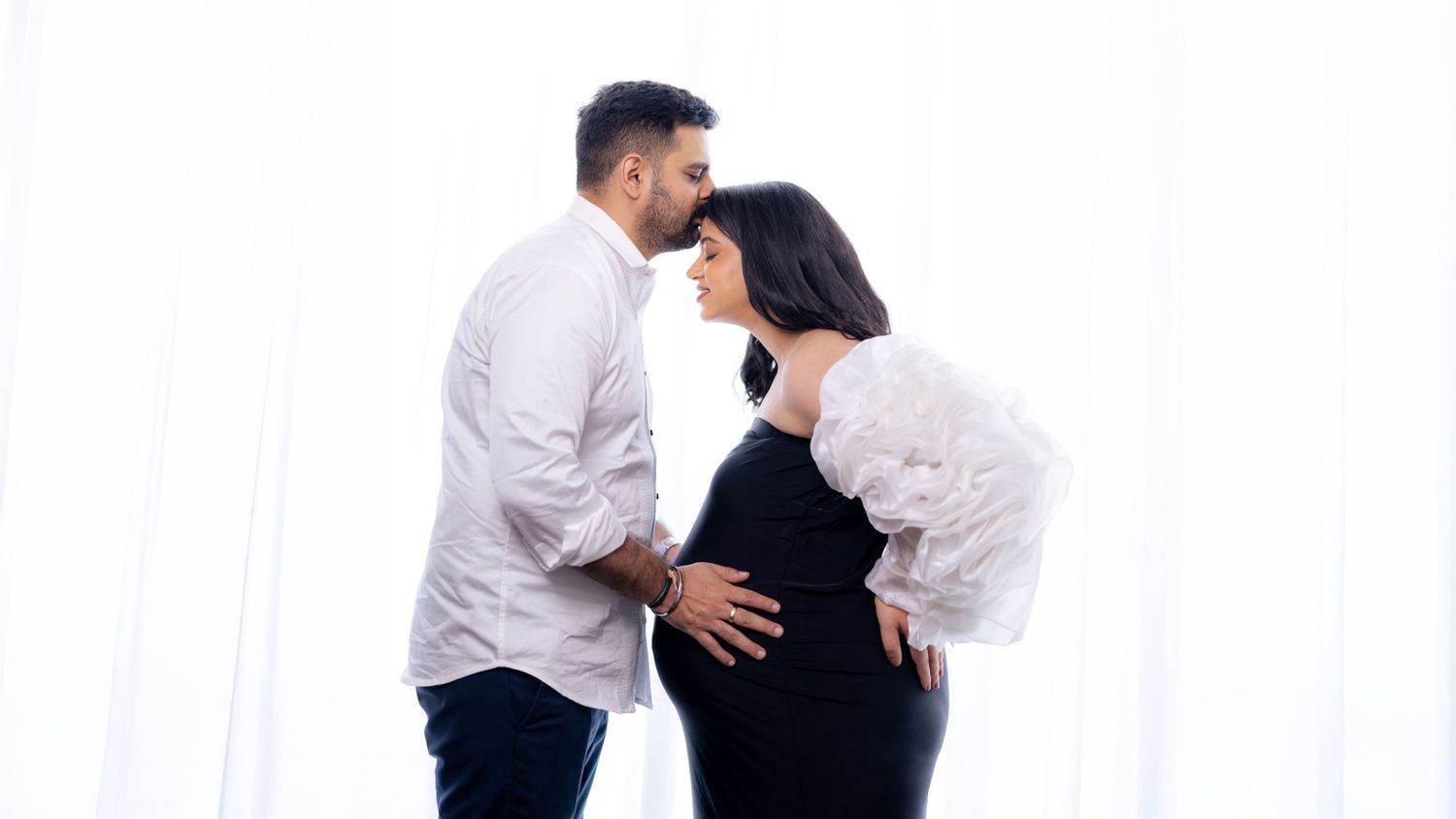 Couple sharing a tender moment during a maternity photo shoot against a white curtain backdrop.