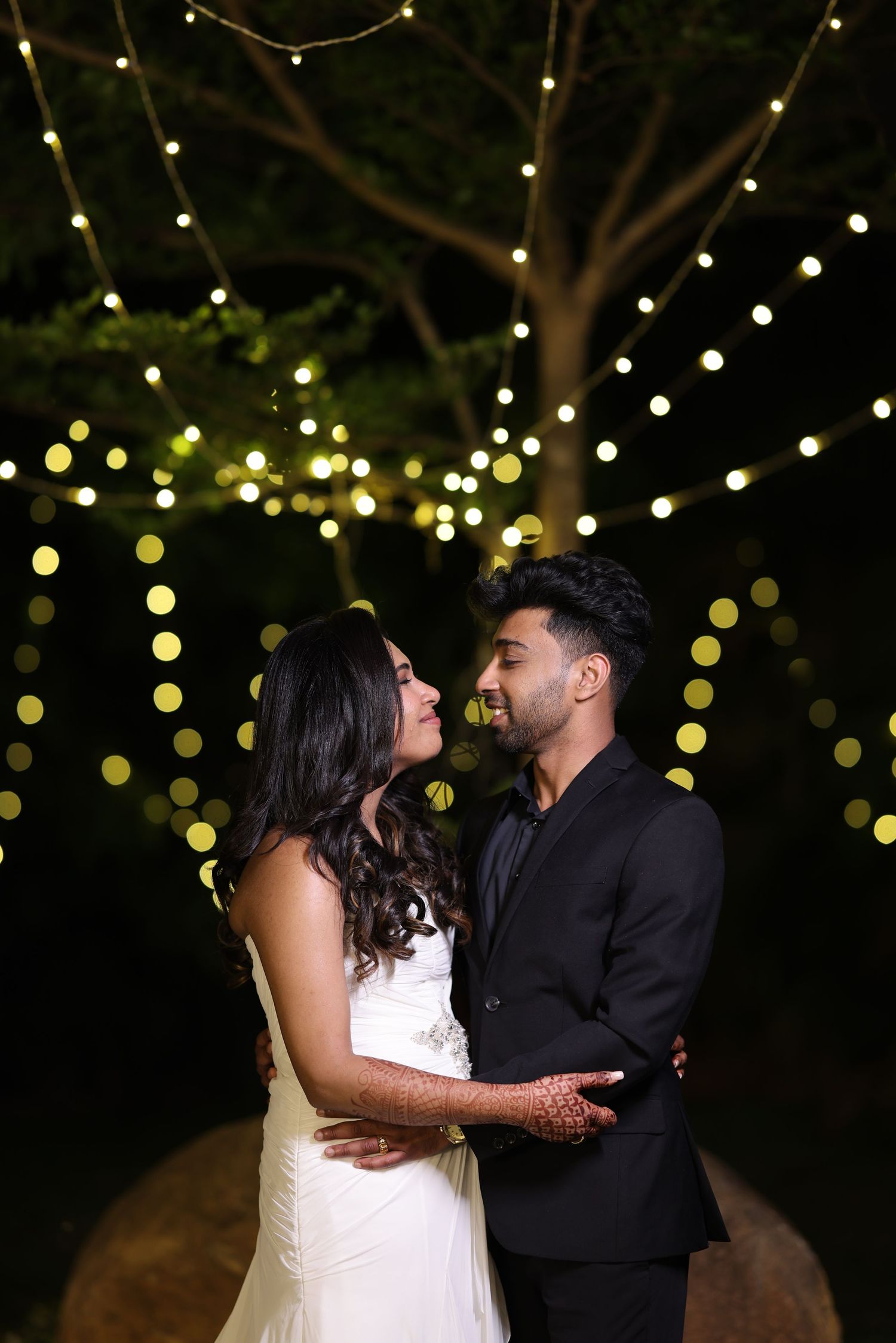 Couple embracing romantically at night under glowing string lights in trees, woman in white dress, man in black suit.