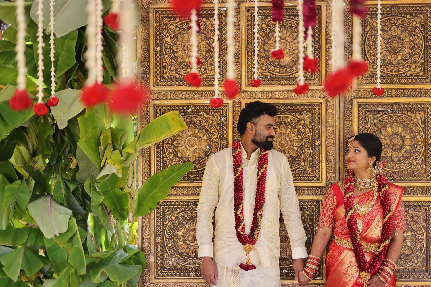 Indian couple in traditional wedding attire with red garlands, standing before ornate golden backdrop with floral decorations.