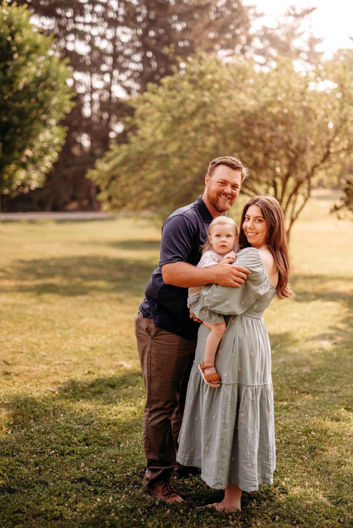 A family poses together outdoors in a sunlit park setting with golden hour lighting and trees in the background at Horticulture Park