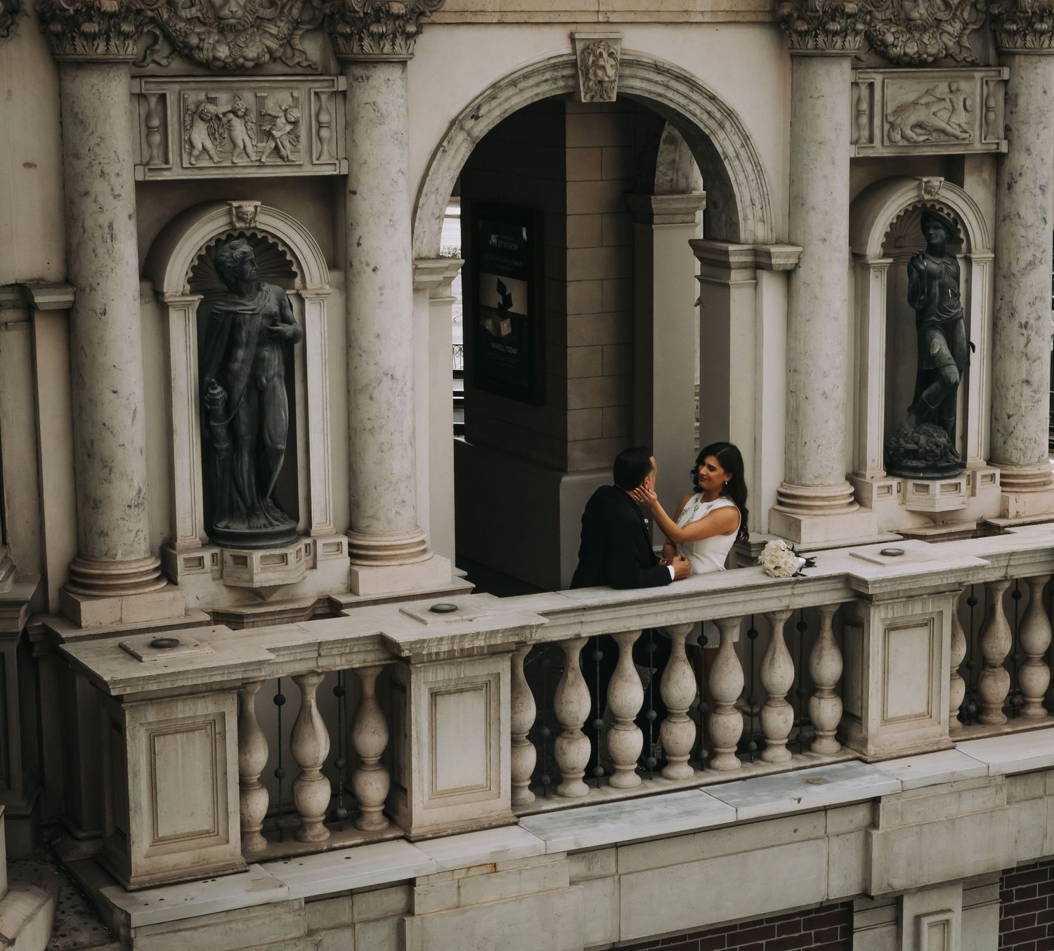 Newlyweds sharing a romantic moment on an ornate balcony with classical statues and balustrades.
