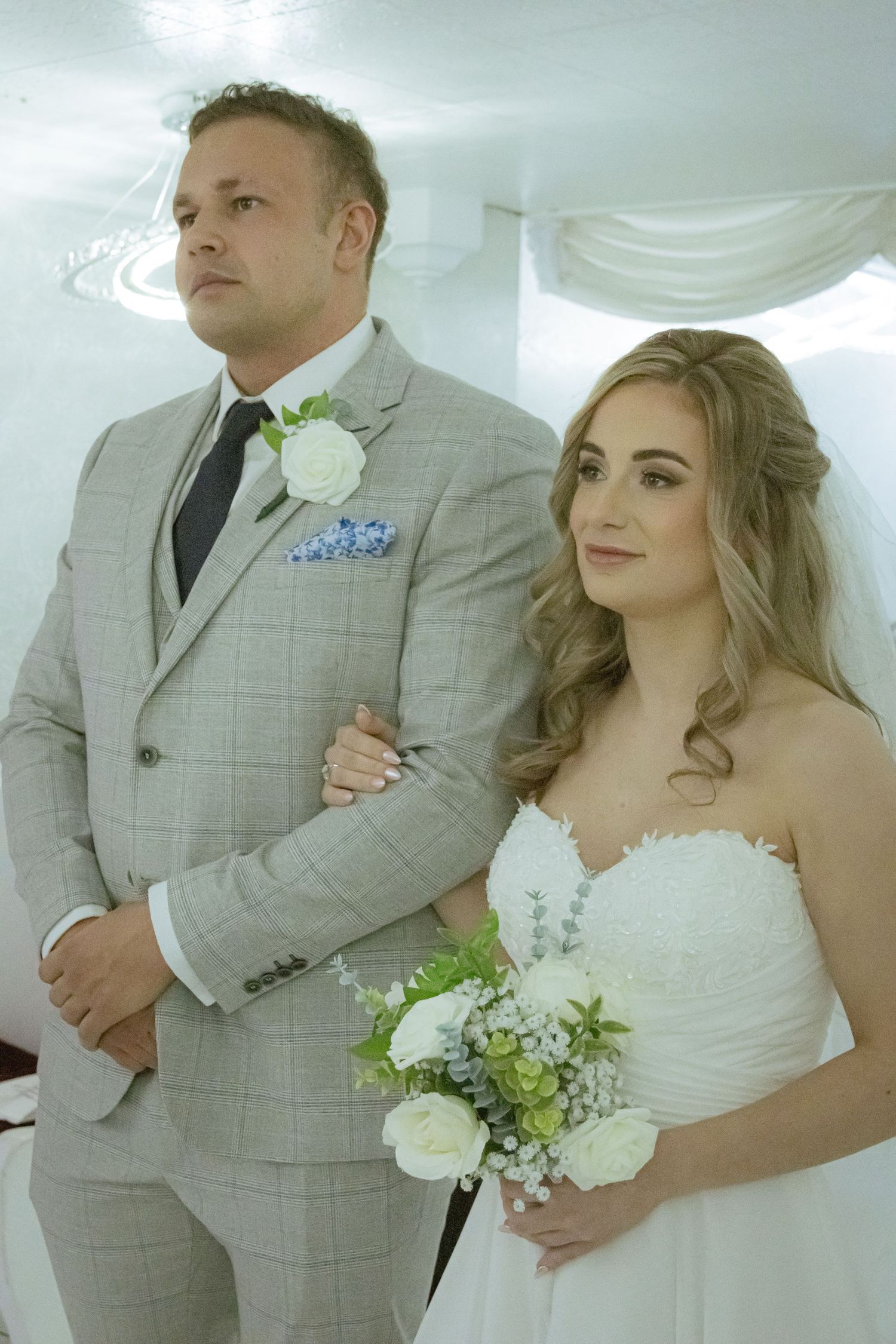 Bride holding white bouquet and groom in grey plaid suit standing together during wedding ceremony.