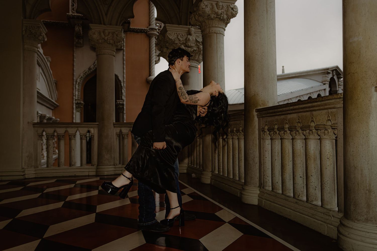 Couple sharing a romantic dip pose in an elegant columned ballroom with checkered marble floors.