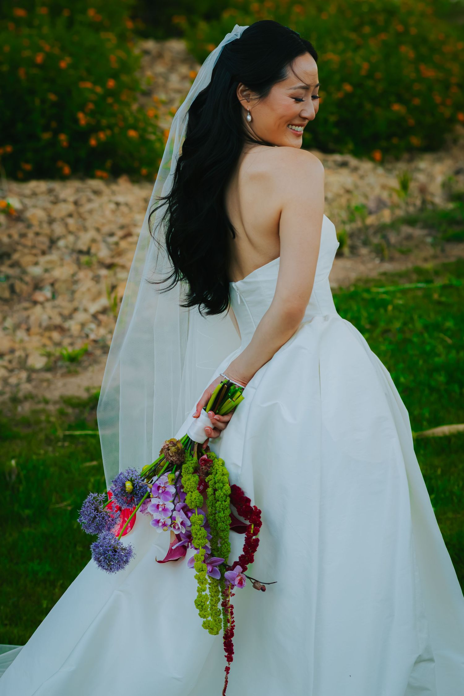 Smiling Asian bride in strapless white gown holding colorful cascading bouquet in garden setting.