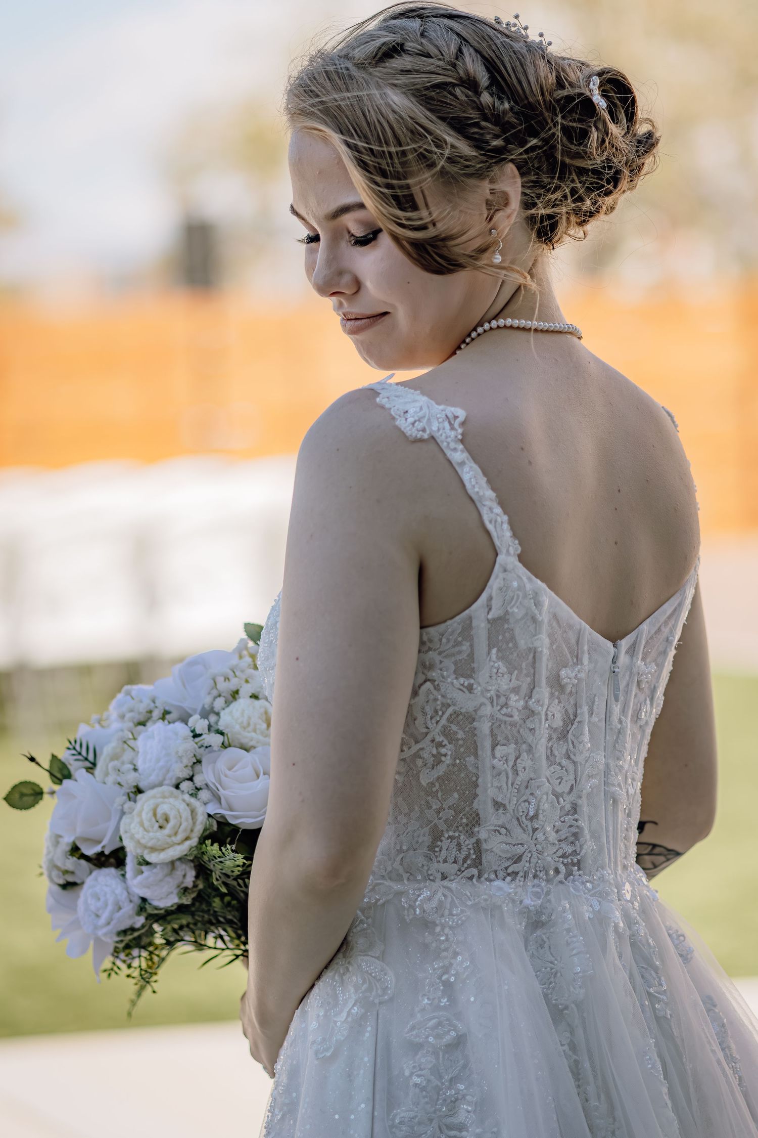 Elegant bride in lace gown viewed from behind, holding white rose bouquet in outdoor setting.