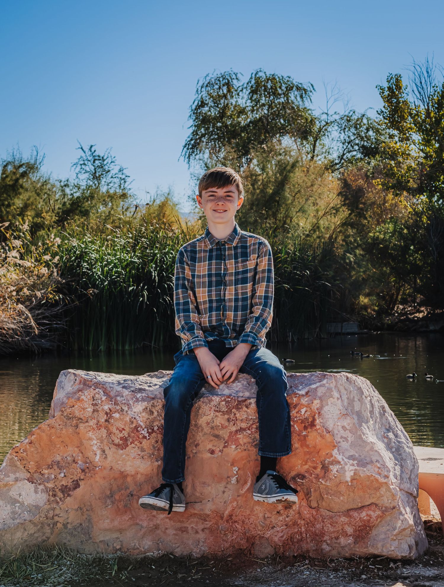 A smiling teenage boy in a plaid shirt sits on a large rock by a peaceful pond.