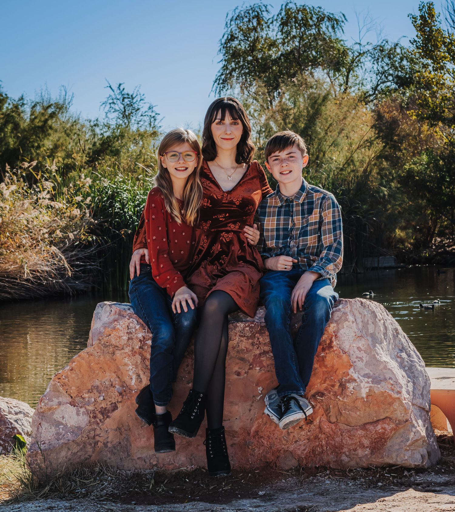 A woman sits on a rock by a pond with a boy and girl on either side of her.