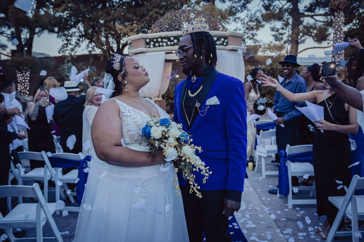 Newlyweds kissing at outdoor ceremony as guests toss white confetti in blue-themed wedding.