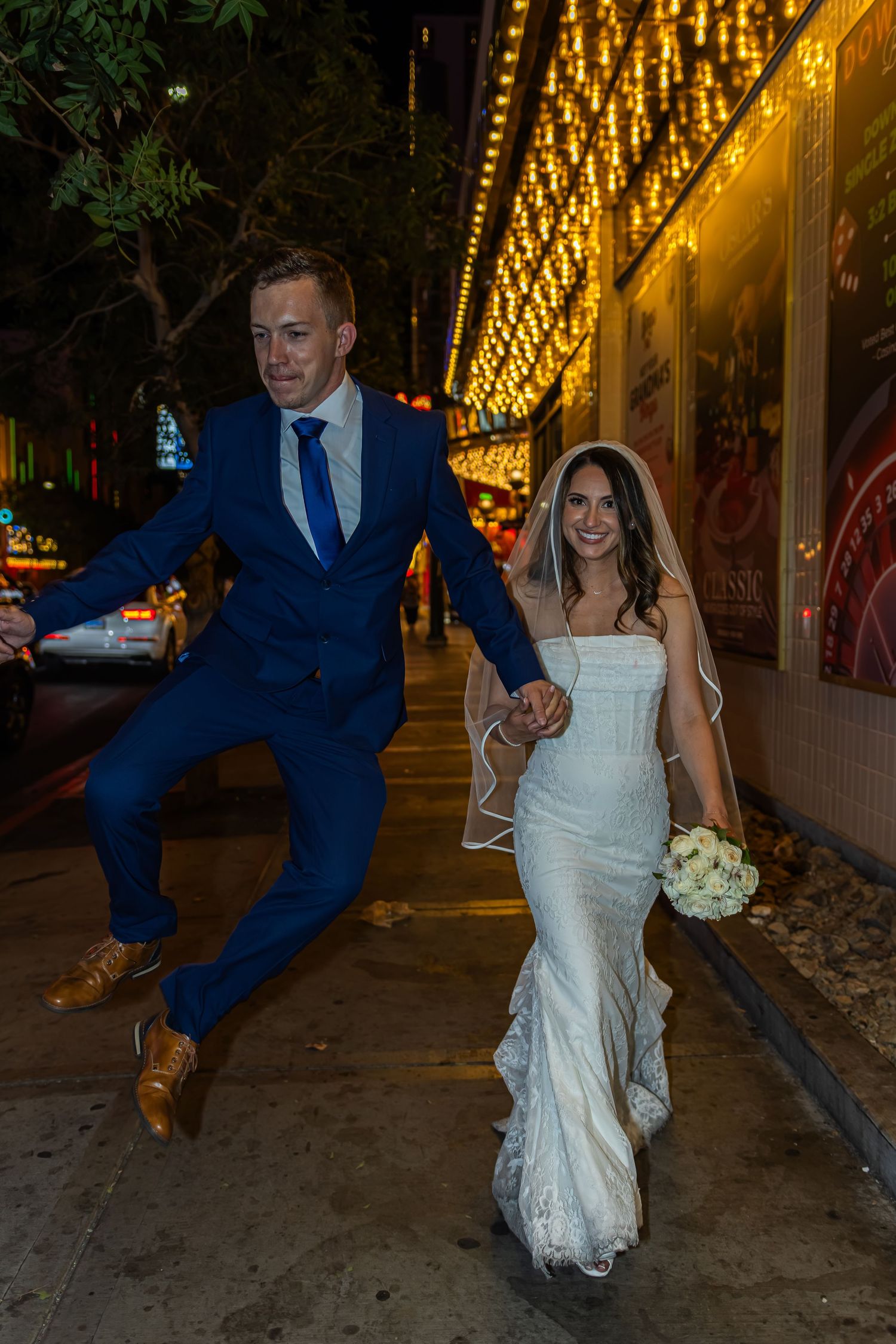 Joyful groom jumps while holding bride's hand outside a neon-lit venue on their wedding night.
