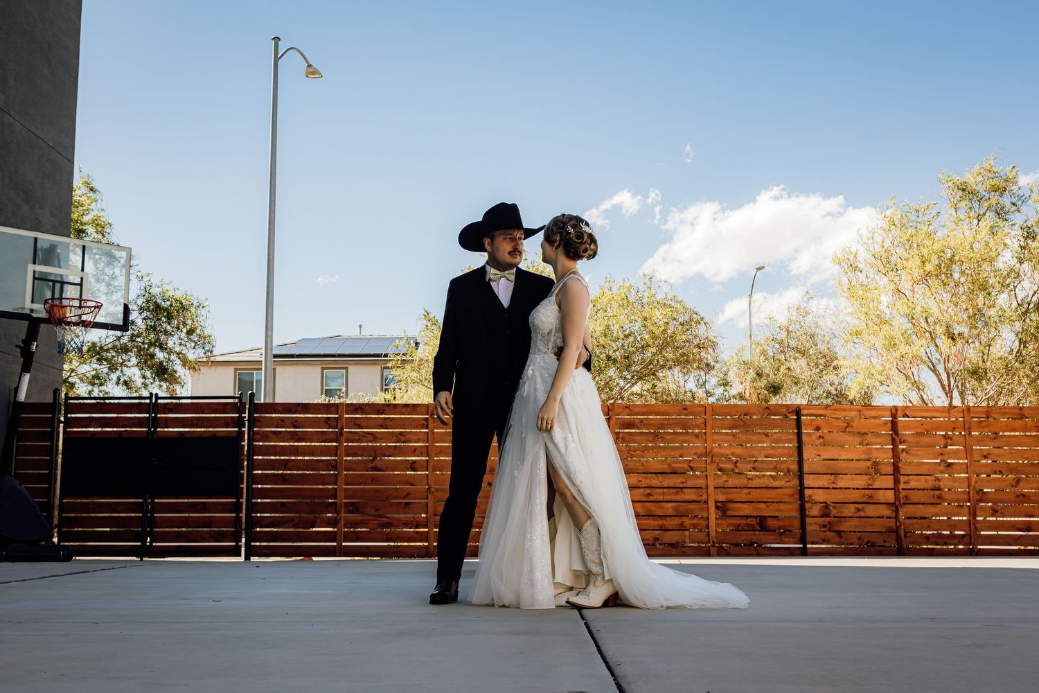 Bride and groom share romantic moment on outdoor court, groom in black cowboy hat and suit.