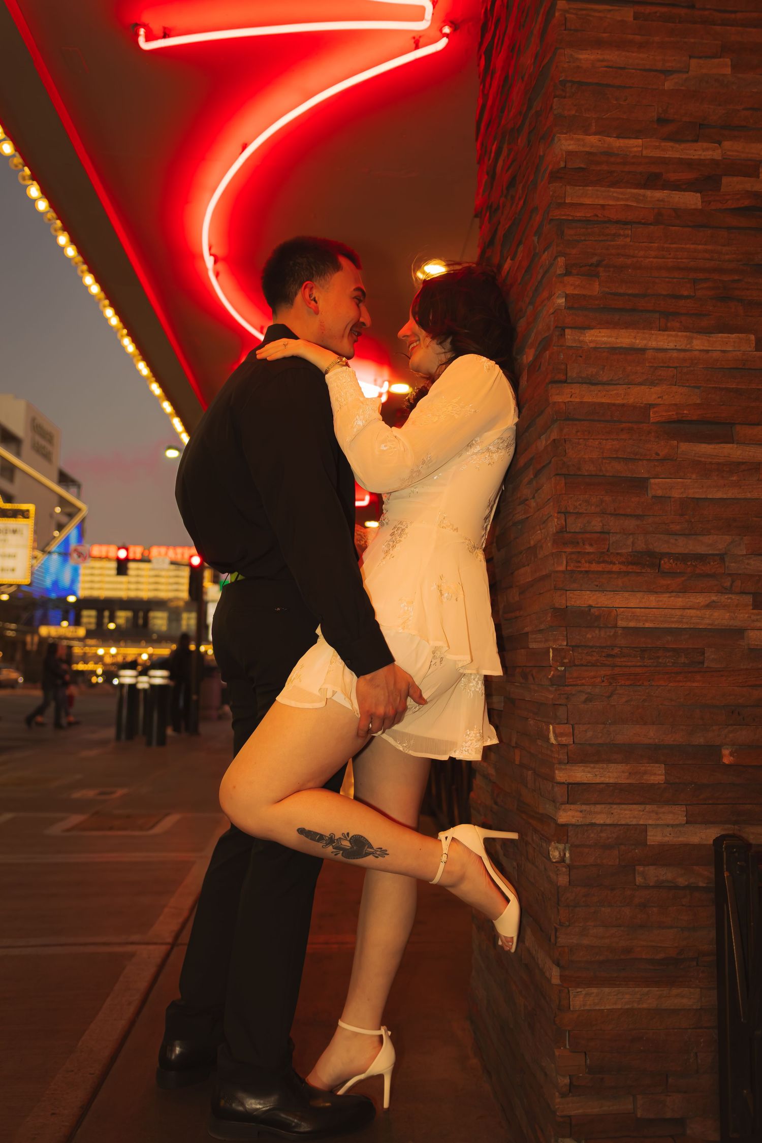 Couple embracing romantically against a brick wall with red neon lights, woman in white dress lifting her leg.