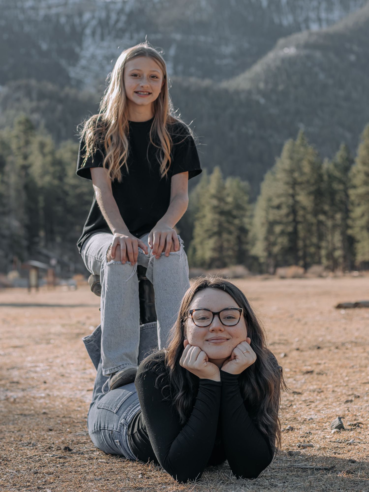 Two girls pose outdoors with mountains and pine trees, one standing, one lying down.