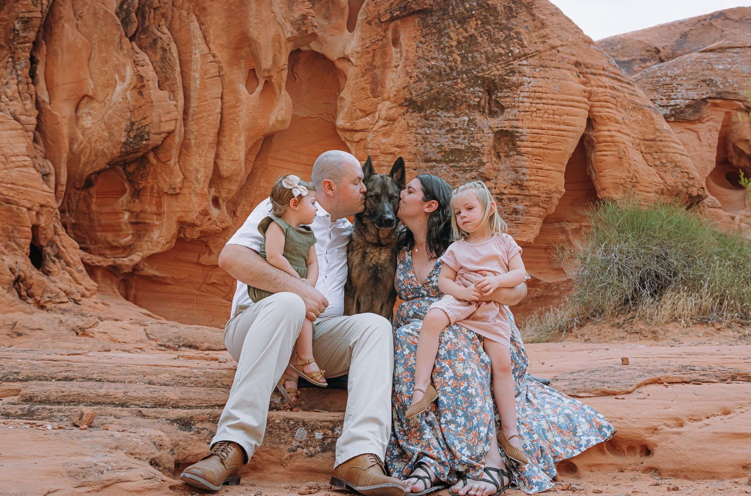Couple kissing while seated on red rock with a German Shepherd and young child in a scenic desert canyon.