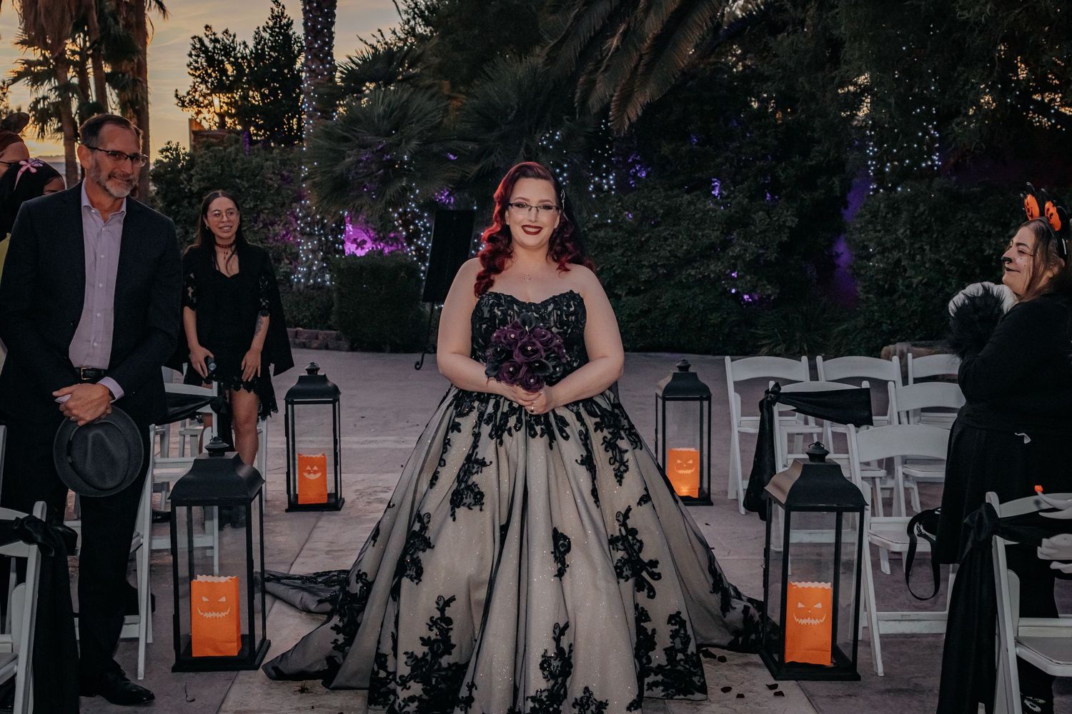 Bride walks down aisle in black and gold gown past lanterns at outdoor evening wedding ceremony.