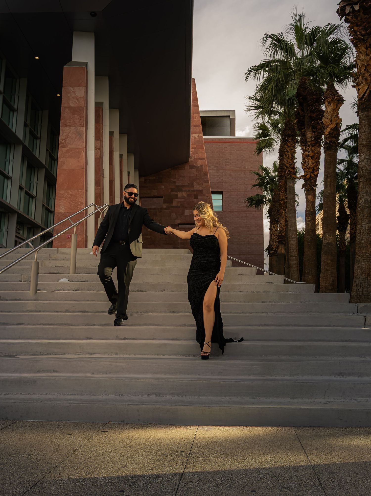 Elegant couple in black formal wear descending outdoor stairs with palm trees at sunset.