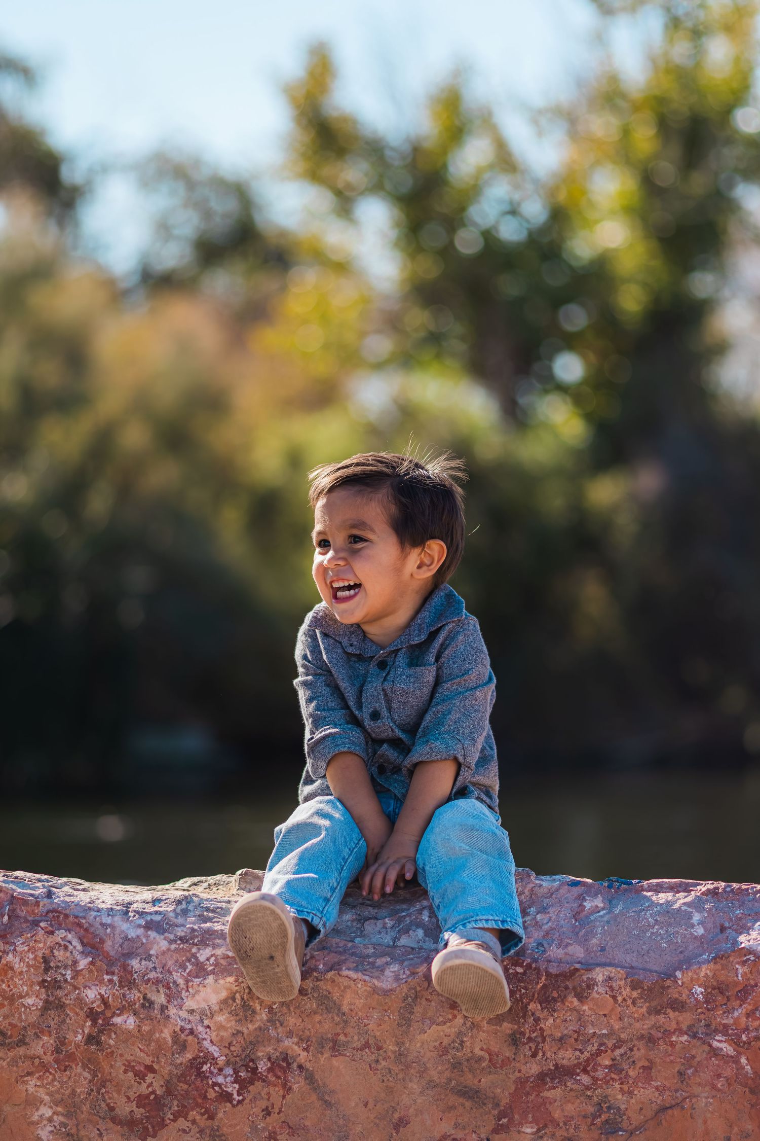 Happy toddler boy sitting on a rock laughing outdoors on a sunny day.