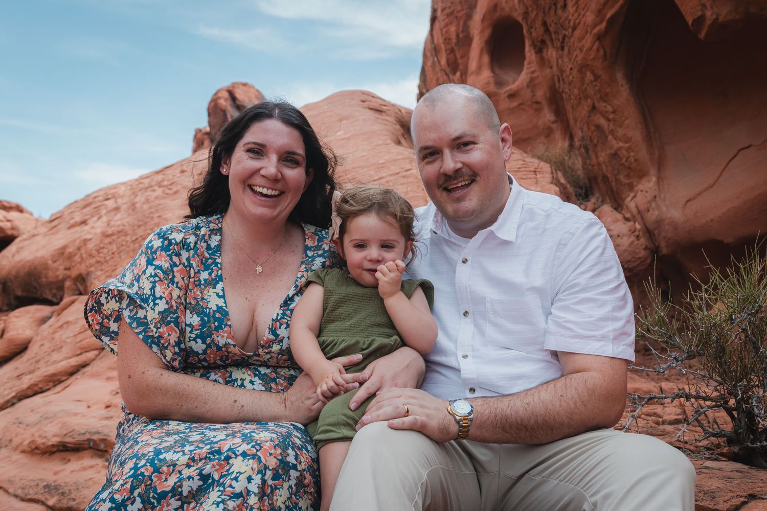Smiling couple with their toddler sitting together against red sandstone rock formations.