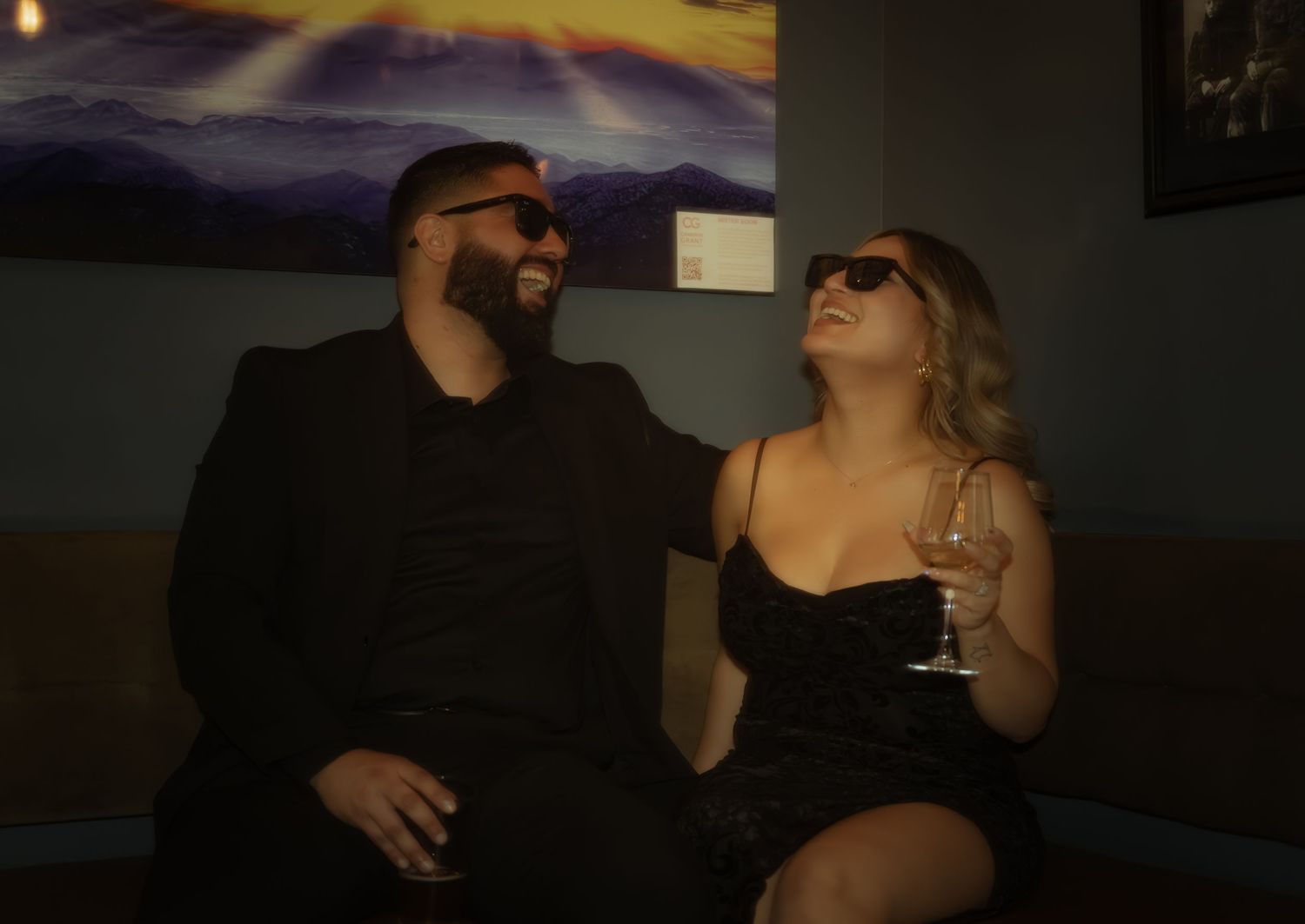 Stylish couple in black outfits wearing sunglasses sharing drinks on a lounge sofa.