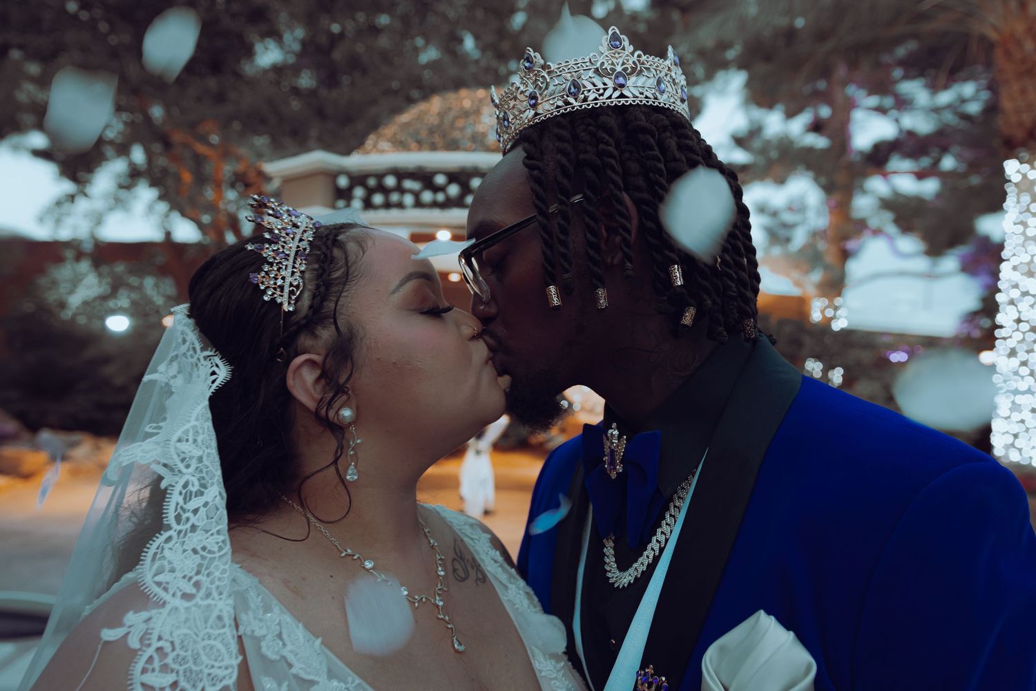 Bride and groom sharing romantic kiss wearing crystal crown and blue suit amid falling confetti.