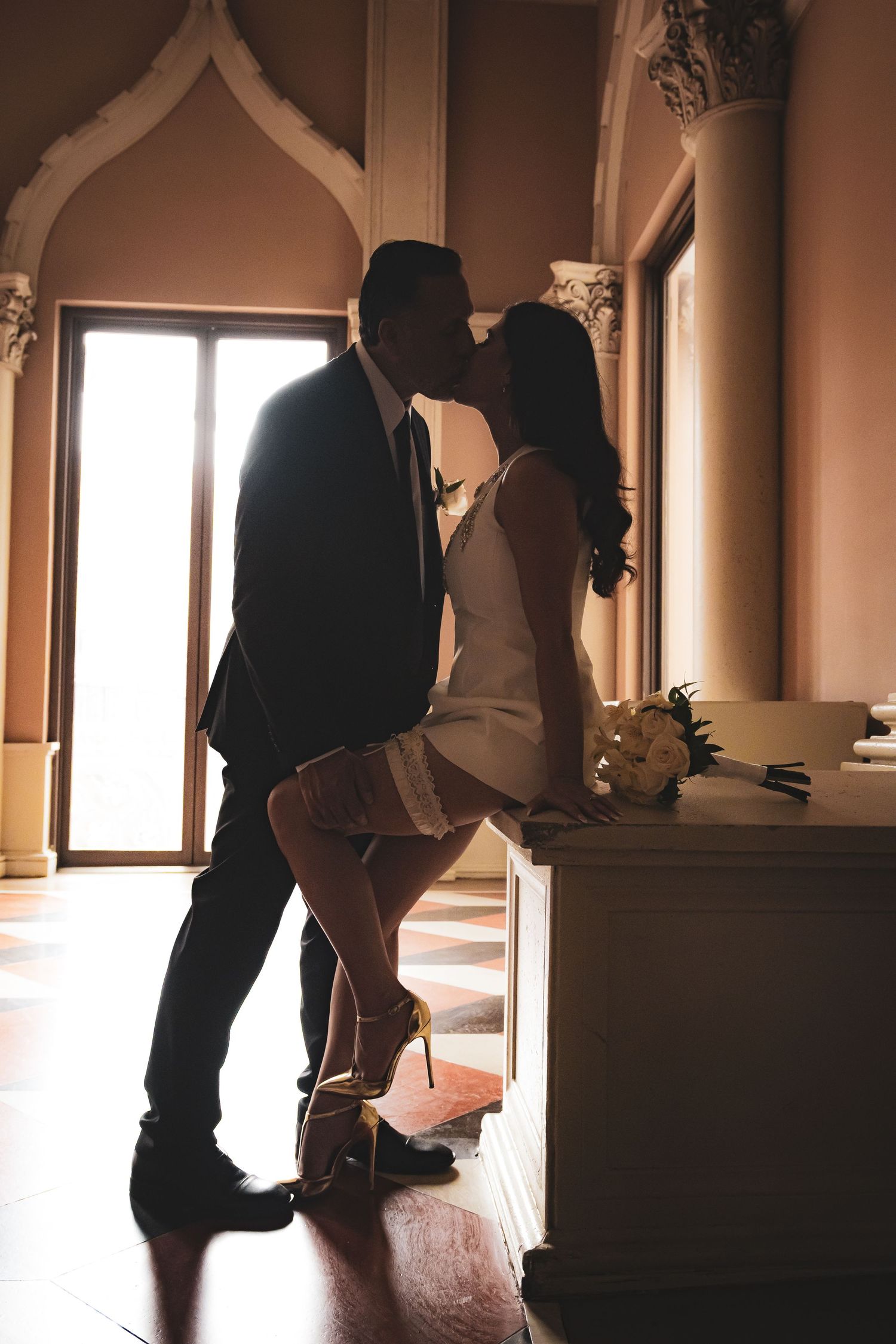 Couple sharing a passionate kiss in silhouette inside elegant venue with arched windows and marble decor.