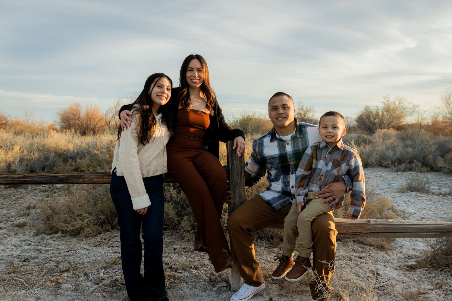 Family of four smiling together outdoors on rustic fence rails with desert shrubs background.