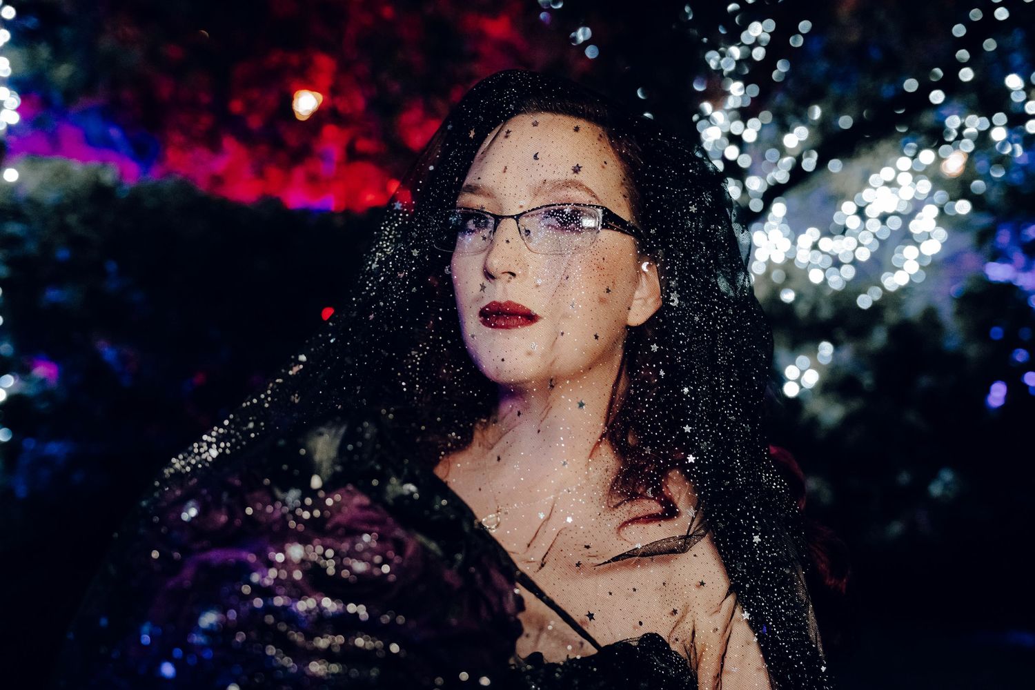 Woman in black lace dress with glasses poses dramatically among colorful bokeh lights at night.