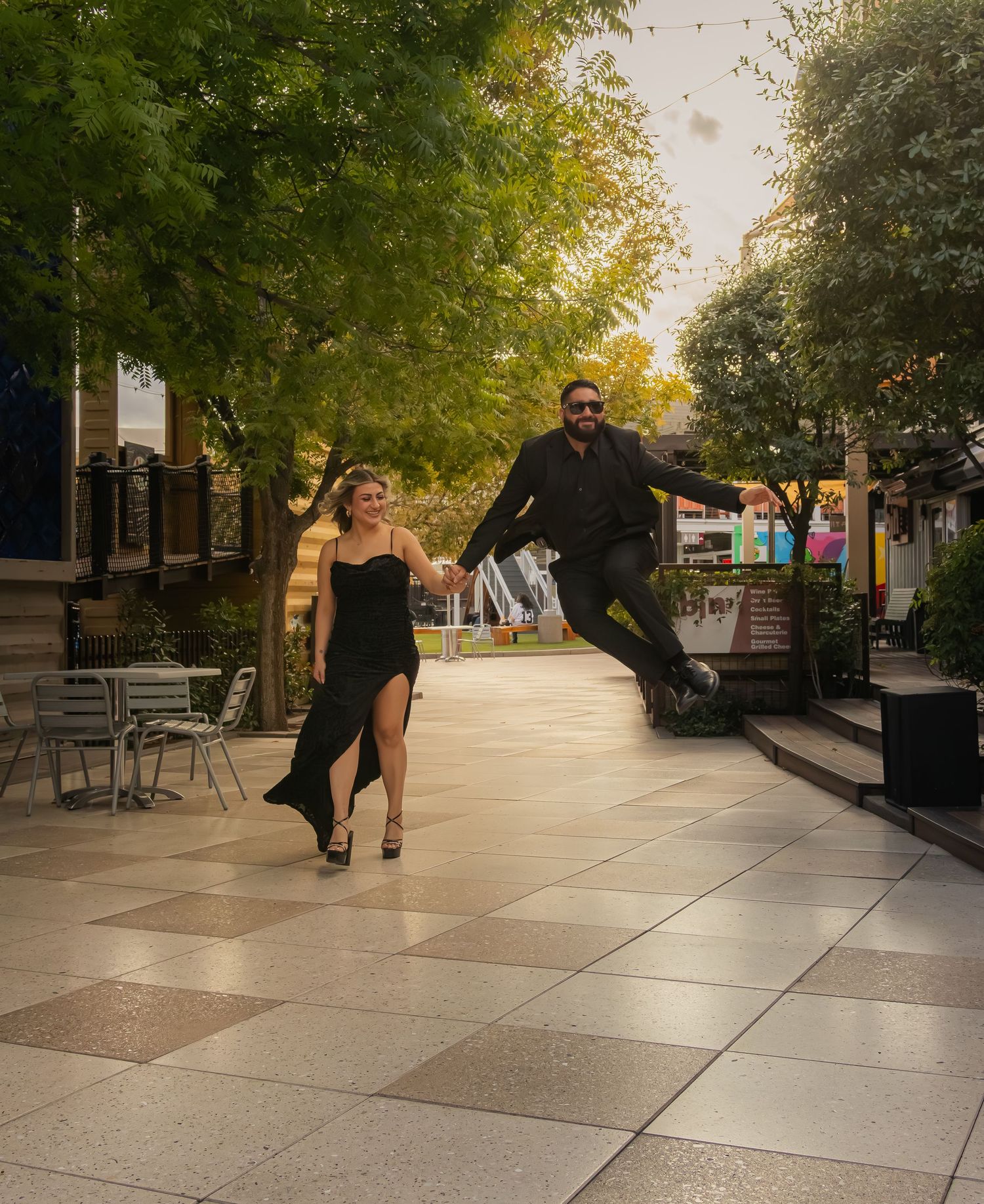 Joyful couple walking hand-in-hand as man leaps in the air along a tree-lined plaza.