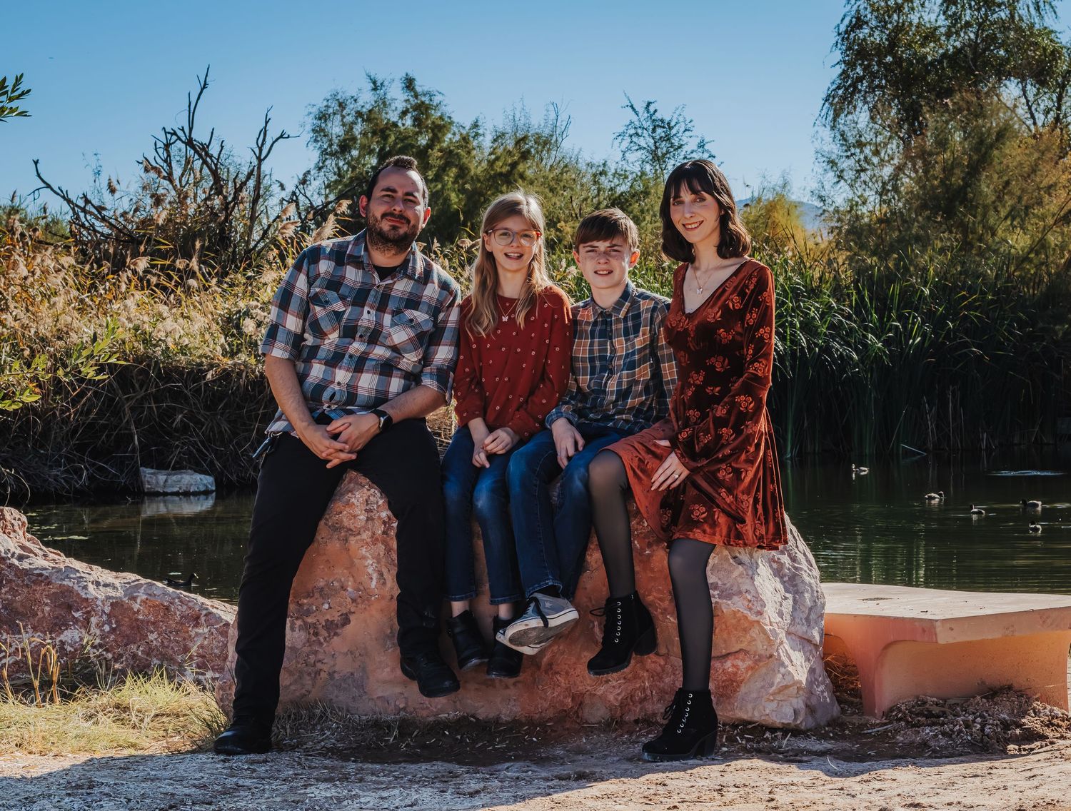 A family of four sits together on a large rock beside a tree-lined pond outdoors.
