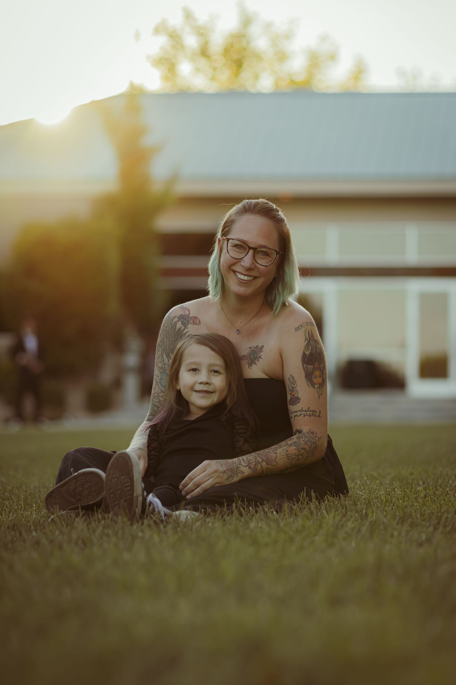 A smiling tattooed mother sits on green grass holding her young daughter close during a golden sunset.