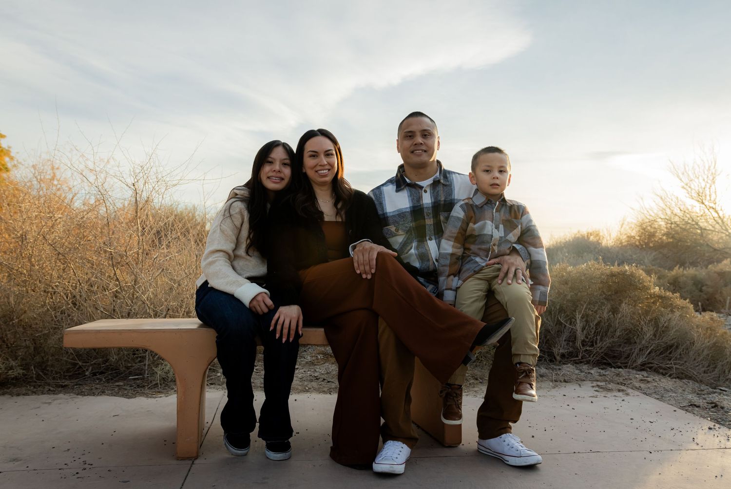 Family of four sitting together on a bench outdoors with desert shrubs and a soft cloudy sky behind them.