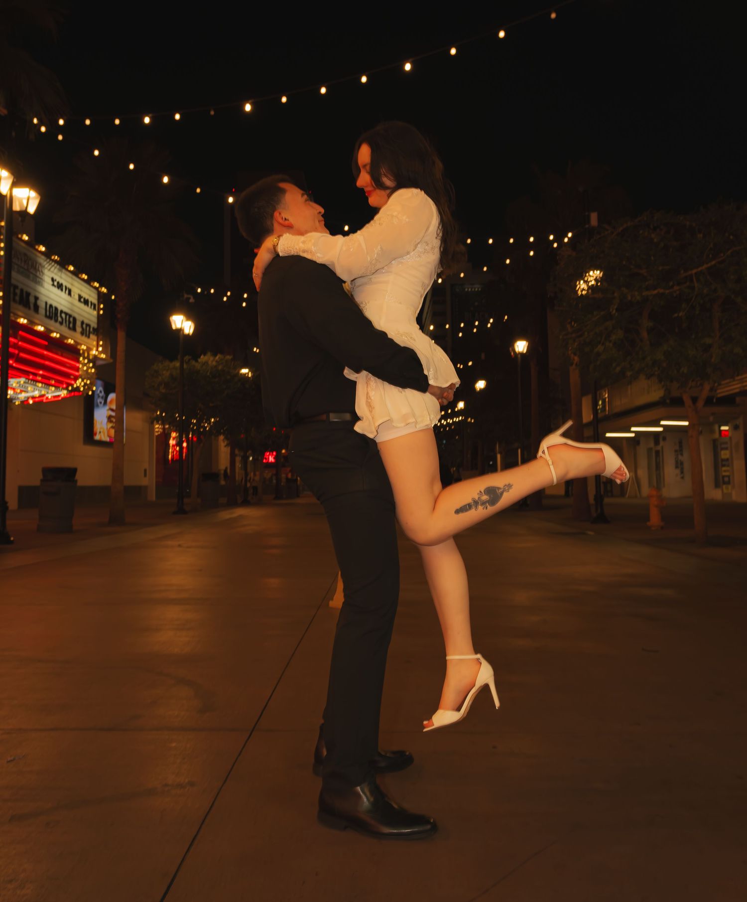 Man lifting woman in white dress and heels under string lights on a romantic Las Vegas night street.